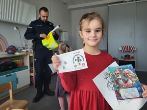 dziewczynka trzymająca w ręku  paszport z odbitymi liniami palilarnymi, ulotke i kolorowankę, za nia w tle policjant wyjmujący odblaski z worka