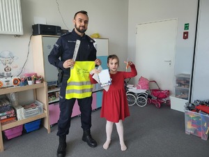 dziewczynka trzymająca w ręku ulotke i kolorowankę, obok policjant z kamizelka odblaskową w rękch