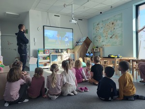 umundurowany policjant stojący przed siedzącymi na podłodze przedszkolakami, za policjantem wyświetlona bajka.