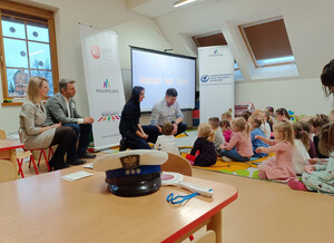 policjantka z wicemarszałkiem kucają przed siedzącymi na podłodze przedszkolakami, za nimi wyświetlona prezentacja, obok wójt i kobieta siedzą na ławce