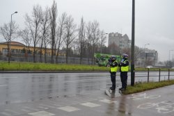 na zdjęciu dwóch policjantów ruchu drogowego, jeden trzyma w ręku ręczny miernik prędkości tzw. radar oraz tarczę do zatrzymywania pojazdów. W tle jezdnia i jadące pojazdy, pas zieleni oraz budynki.