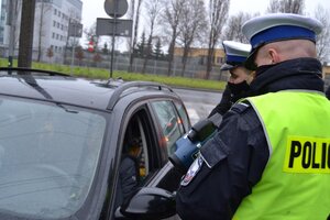 na zdjęciu dwóch policjantów ruchu drogowego, jeden trzyma w ręku ręczny miernik prędkości tzw. radar stoją przy zatrzymanym do kontroli drogowej pojeździe.