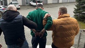 Funkcjonariusze operacyjni doprowadzają zatrzymanego, który ma założone kajdanki na ręce trzymane z tyłu
