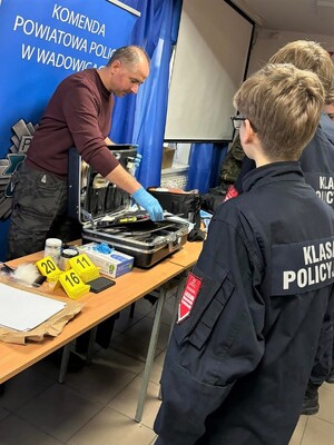 Technik kryminalistyki prezentuje swój sprzęt, przed nim uczniowie w mundurach. W tle policyjny baner z napisem Komenda Powiatowa Policji w Wadowicach.
