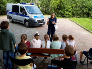 Policjantka prowadzi prelekcję dla dzieci, w tle radiowóz policyjny