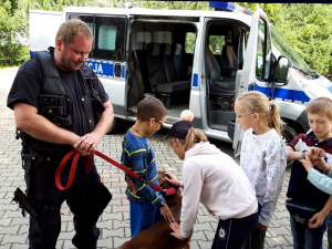 przy radiowozie grupa dzieci rozmawia z przewodnikiem psa służbowego, dzieci głaskają policyjnego psa