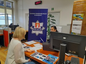 policjantka opowiadająca o pracy siedzącej po przeciwnej stronie biurka kobiecie, za policjantką baner KPP Kraków, na biurku ulotki
