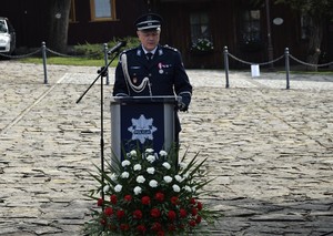 Komendant Powiatowy Policji w Wadowicach przemawia. Stoi za mównicą na której jest logo Policji. Przed nią bukiet biało czerwonych kwiatów.